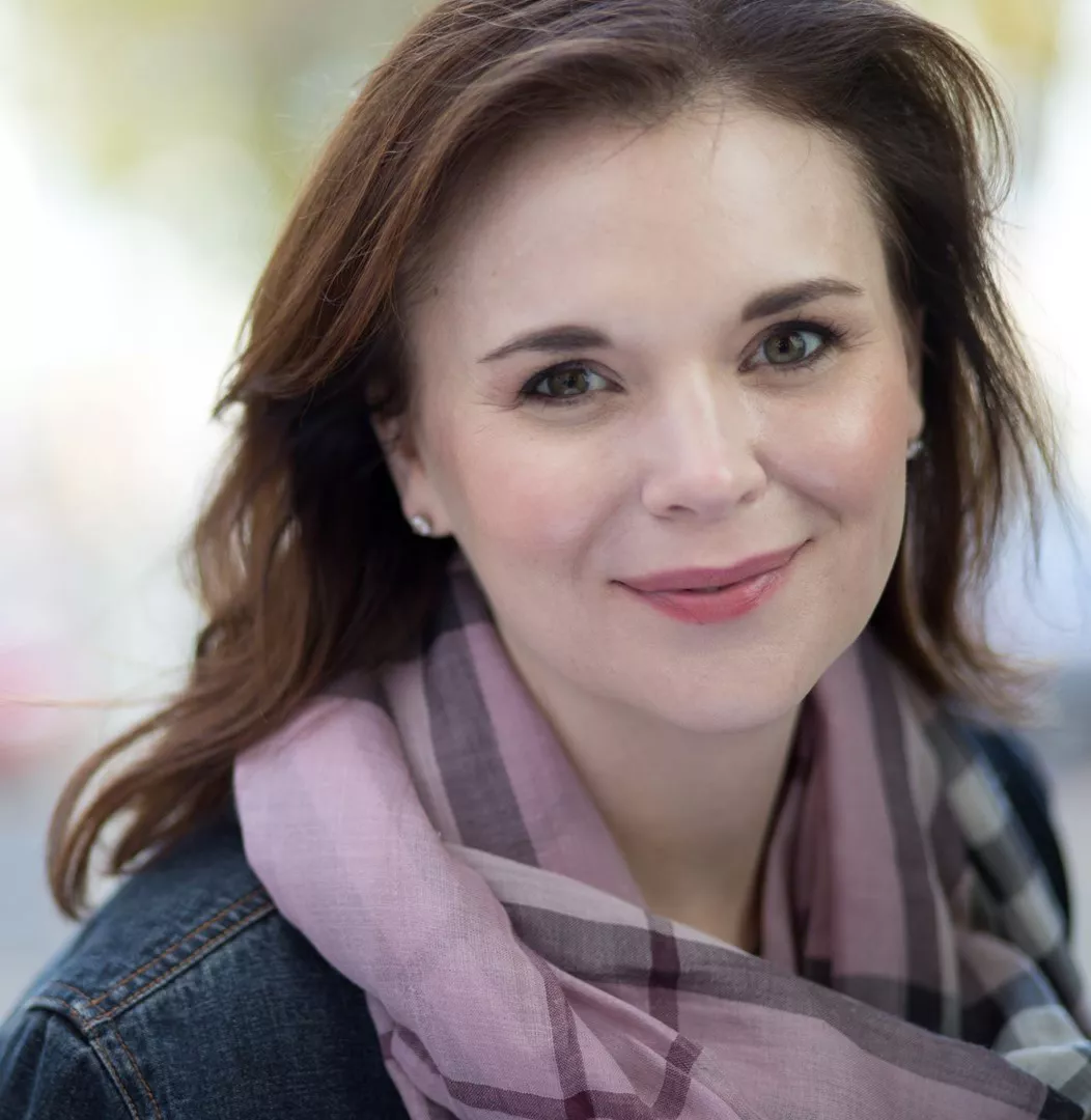 A woman with brown hair is seen wearing a pink plaid scarf and a denim jacket. The setting appears to be outdoors with a blurred background of trees and light. The scarf features a checkered pattern in soft pink and neutral tones, complementing the casual style.